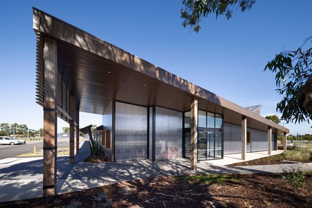 Single-storey contemporary commercial building with a long, low-slung flat roof and deep covered entrance canopy. The facade combines dark metal framing, translucent corrugated panels, extensive clear glazing, and warm brown timber-toned cladding. A paved forecourt and parking area sit alongside sparse landscaped planting.