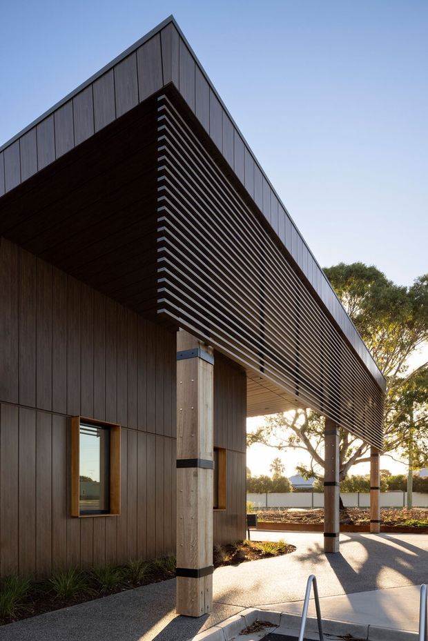 Contemporary single-storey building exterior with dark vertical cladding, a dramatic projecting flat roof and deep covered entry or outdoor area. Slim horizontal louvres form a prominent sun-screen beneath the roof edge, supported by square timber or concrete-look posts with dark metal bands. Small windows, paved paths and landscaped planting are visible, with a clean minimalist composition and warm late-day light.