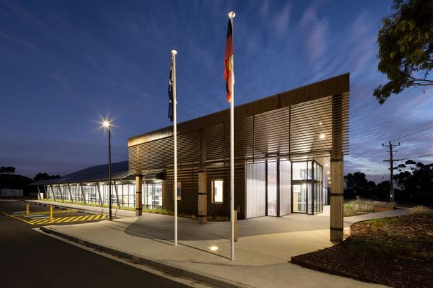 Twilight exterior view of a contemporary institutional or commercial building. The low-rise structure features extensive glass walls, a broad projecting entrance canopy with horizontal timber-toned slatted screening, dark vertical cladding, illuminated interiors, flagpoles, paved access paths, and a covered glazed walkway extending to the left.