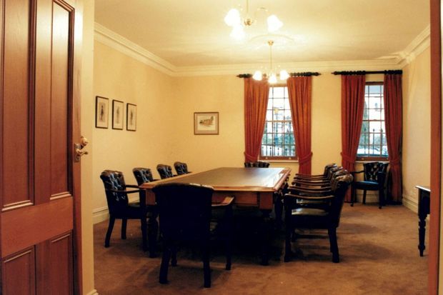 A formal dining room with a large rectangular timber dining table surrounded by dark upholstered chairs. The room has warm beige walls, carpeted flooring, ornate white cornices and ceiling detailing, tall multi-pane windows with deep red drapes, framed artwork, and a traditional central chandelier. A paneled timber door is visible at the left.