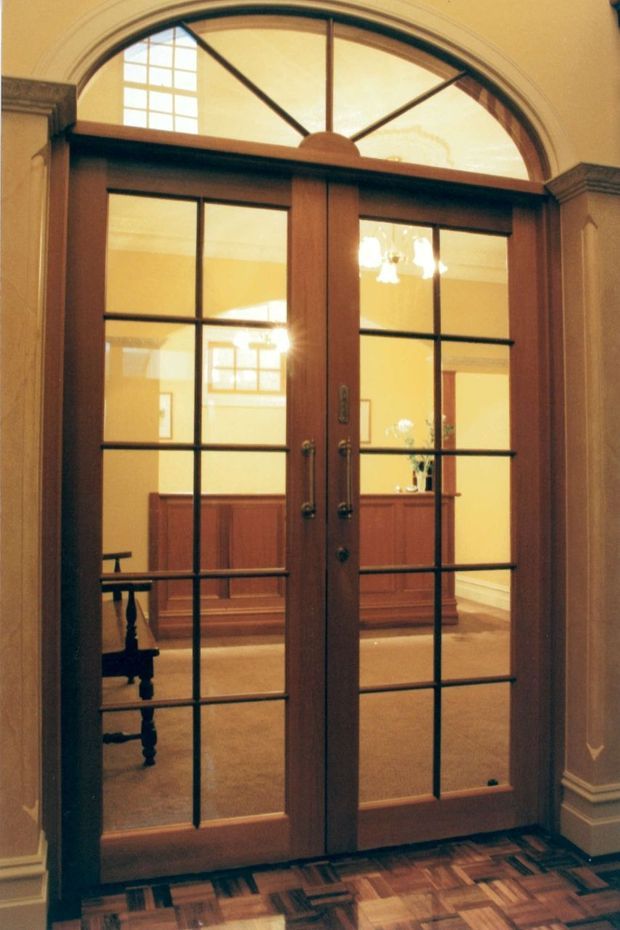 Interior view of a traditional entry or hallway focused on a pair of dark timber-framed glazed double doors with black muntins and an arched fanlight above. The doors open toward a warm, carpeted room with dark timber wall panelling, a console or cabinet, chandelier lighting, and a partially visible traditional chair. The surrounding walls feature ornate moulded trim and wainscoting, while the foreground has patterned parquet timber flooring.