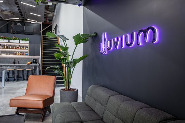 A stylish commercial lounge and reception area with a dark charcoal feature wall, purple neon signage, a grey upholstered sofa, and a tan leather lounge chair. Indoor plants add greenery, while an open bar or kitchenette area with shelving and stools is visible in the background. The space has exposed ceiling services, linear lighting, and a contemporary industrial fit-out.
