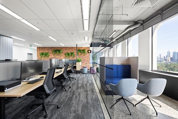 Bright contemporary open-plan office interior with rows of workstations, black ergonomic office chairs, large monitors, a carpeted circulation zone, and a small informal seating area beside full-height windows. Exposed services and a brick feature wall with planted greenery add visual interest, while the windows overlook a dense city skyline.