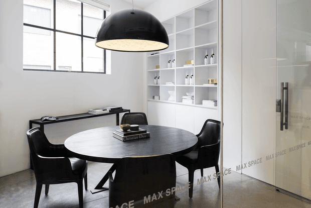 Bright contemporary dining area with a round dark dining table surrounded by black sculptural chairs. A large black dome pendant light hangs above, while white built-in open shelving displays tableware and decorative objects. Large industrial-style framed windows, a slim console, polished concrete flooring, and a glazed door create a clean, refined interior.