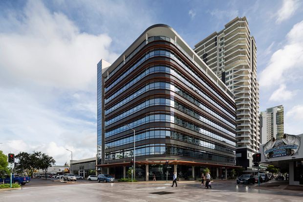 Street-level exterior view of a contemporary multi-storey commercial or mixed-use building on a prominent urban corner. The main building has rounded corners, extensive horizontal bands of dark reflective glazing, reddish-brown cladding accents, and a curved rooftop element. Taller residential or office towers rise behind it, with traffic lights, sidewalks, storefronts, and pedestrians at street level.
