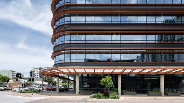 Exterior street-level view of a contemporary multi-storey commercial building with rounded glazed corners, horizontal dark metal and warm timber-toned bands, a projecting covered entrance canopy, and ground-floor retail or office glazing. Urban streetscape and neighboring buildings are visible around the main facade.