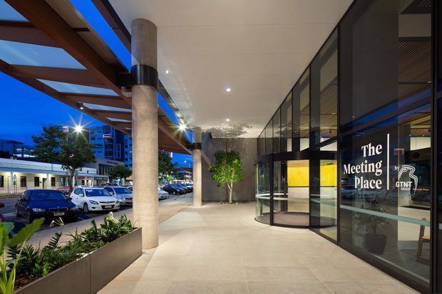 A dusk exterior view of a contemporary commercial building entrance. A wide covered pedestrian forecourt features large round concrete columns, timber-toned overhead beams, recessed lighting, tiled paving, and landscaped planter beds. Full-height dark-framed glazing encloses the entrance, with revolving and hinged glass doors and signage reading “The Meeting Place” and “GTNT.” Urban buildings, parked cars, and a street are visible beyond the covered walkway.