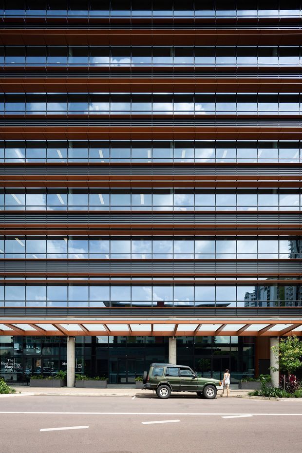 Street-level view of a contemporary multi-storey commercial building with a highly glazed facade, repeated horizontal timber-toned sun-shading fins, dark metal framing, and a projecting entrance canopy. The ground floor contains glazed lobby and retail or hospitality spaces, with landscaped planters along the frontage. Reflections of sky and nearby buildings are visible in the curtain wall glazing.