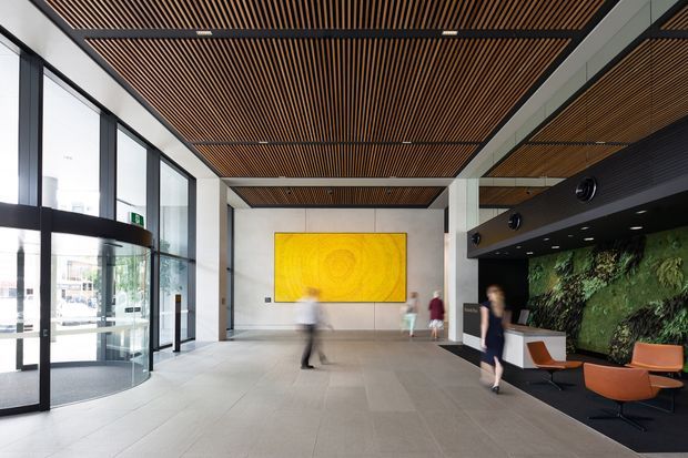 A spacious contemporary commercial lobby or reception area with full-height glass entry doors, pale stone or concrete flooring, white structural columns, and a striking linear timber-slat ceiling. A reception desk with a lush green living wall occupies the right side, while a large abstract yellow artwork forms a focal point at the rear. The space has a refined, modern corporate character with abundant daylight and blurred visitors moving through the lobby.