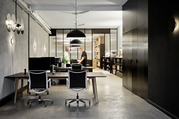 A sophisticated open-plan home office or studio with a long dark worktable, ergonomic swivel chairs, pendant lighting, concrete-style walls and polished concrete flooring. Floor-to-ceiling black-framed glazing separates the workspace from a secondary living or library area with extensive shelving and cabinetry. The space has a refined industrial-contemporary character with a neutral grey, black and white palette.