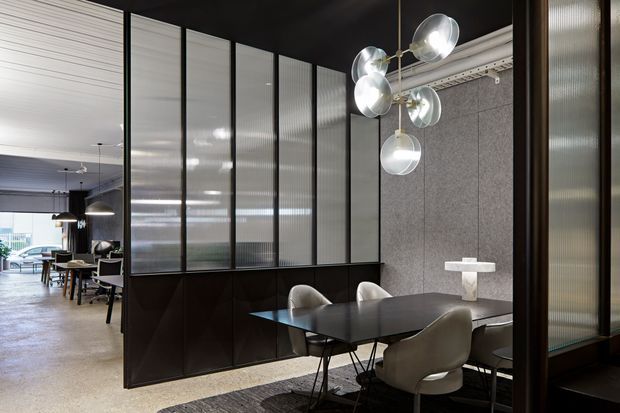 A sophisticated open-plan dining area screened by black-framed translucent ribbed-glass partitions. A dark rectangular dining table is surrounded by upholstered chairs, with a sculptural cluster of spherical glass pendant lights overhead. The space features dark cabinetry or partition bases, a grey textured feature wall, pale flooring, and a visible adjoining lounge or work area beyond.