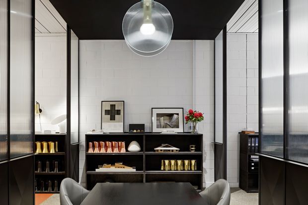 A refined, dark-toned interior featuring a black dining or meeting table with sculptural chairs, a low black display cabinet, framed artwork, books, and decorative objects. White painted masonry walls contrast with black ceiling and slender black-framed translucent partitions. A large spherical pendant light hangs centrally, creating a polished, gallery-like atmosphere.