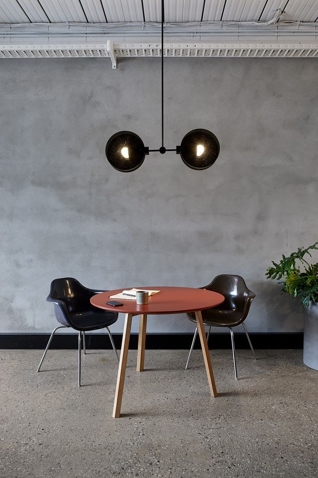 A compact industrial-style dining or meeting nook with a round reddish timber table, two dark molded chairs, and a sculptural black pendant light. The space has a mottled grey concrete-like wall, polished concrete or terrazzo flooring, exposed white ribbed ceiling panels, visible cable trays, and a large indoor plant in a grey planter.