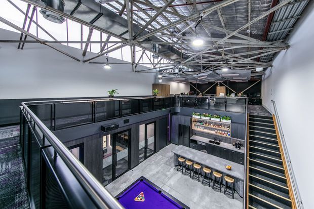 A spacious industrial-style commercial interior viewed from an upper mezzanine. The open-plan venue features exposed steel roof trusses and services, black metal balustrades, a central bar with shelving and greenery, a long stone-look counter with stools, polished concrete flooring, and a staircase descending to the lower level. A partial pool table is visible in the foreground.