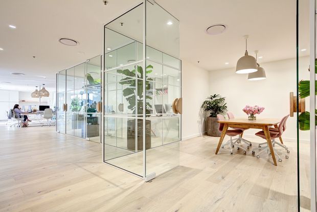 Bright contemporary office interior with a pale timber floor, white walls and ceiling, extensive frameless glass partitions, indoor plants, and an informal meeting area furnished with a timber table, wheeled chairs, and pendant lights. Open workspaces are visible beyond the glazed meeting rooms.