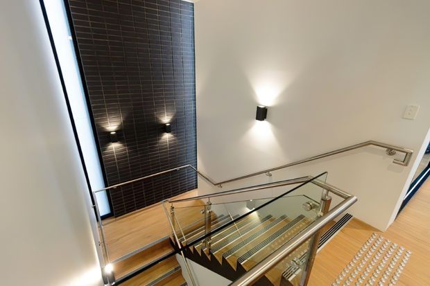 Interior view of a contemporary stairway and landing, featuring timber stair treads, clear glass balustrades with metal handrails, white walls, recessed and wall-mounted lighting, and a tall dark tiled feature wall.