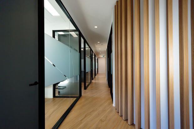 A contemporary office corridor with warm timber-look flooring, black-framed glass meeting rooms on the left, and a striking vertical timber slat feature wall with integrated light strips on the right. Recessed ceiling downlights and glazed partitions create a bright, professional workspace.