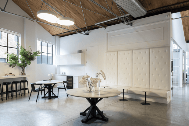 A bright, open-plan commercial-style interior combining a dining and lounge area. The space features polished concrete flooring, white walls and built-in cabinetry, a large tufted cream upholstered wall banquette, round tables, black stools and chairs, exposed timber ceiling boards, visible metal ductwork and suspended track-style services. Large industrial-framed windows provide abundant natural light.
