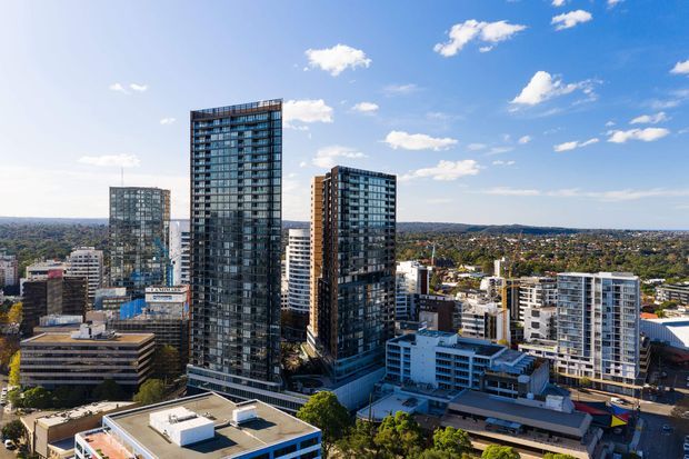 Aerial view of a dense urban precinct featuring several modern high-rise residential or commercial towers, mid-rise buildings, flat roofs, extensive curtain-wall glazing, and a suburban cityscape extending into the background under a bright blue sky.