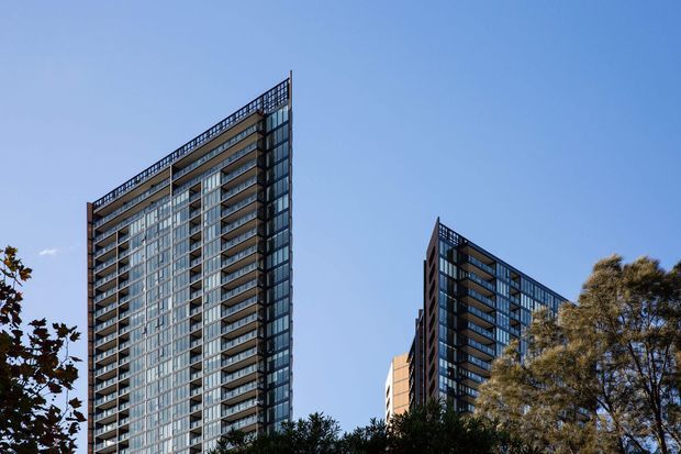 Exterior architectural photograph of two tall contemporary apartment towers with extensive glazed facades, stacked balconies, dark structural framing, and rooftop or parapet railings. Trees partially screen the lower levels, with a clear blue sky dominating the background.