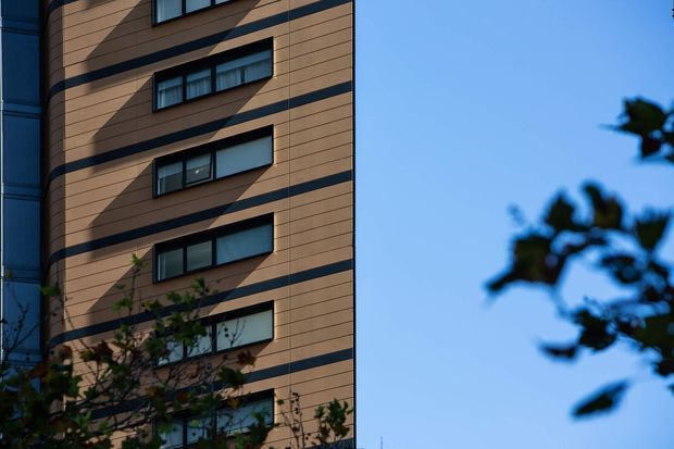 Exterior architectural photograph of a multi-storey building facade with warm brown horizontal cladding, dark horizontal bands, and projecting rectangular windows with dark frames. A narrow blue-grey adjoining facade is visible at left, while blurred tree branches and clear sky occupy the right side.