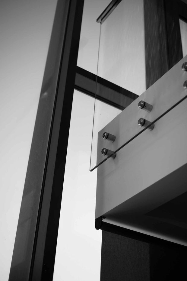 Close-up monochrome architectural detail showing a floating stair or landing edge with clear glass balustrade panels secured by circular metal standoffs. Dark timber or metal framing and a white wall create a strong geometric, high-contrast composition.