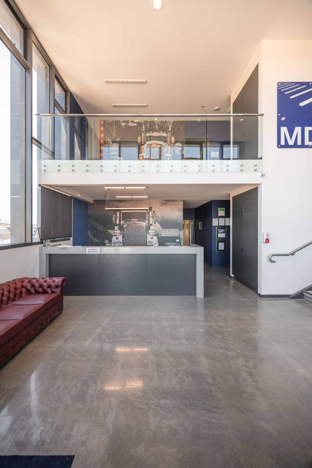 A bright, double-height commercial reception or showroom interior with polished concrete flooring, extensive floor-to-ceiling glazing along the left wall, dark built-in reception cabinetry, and a glass-balustraded mezzanine. Industrial equipment or display machinery is visible behind the reception counter, while a partial leather Chesterfield sofa sits in the foreground. A stair with a metal handrail leads upward on the right.