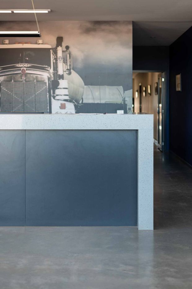 A moody modern interior featuring a large dark blue-grey reception or kitchen island with a pale speckled benchtop. A dramatic black-and-white truck mural forms the backdrop, while a dim hallway with framed artwork extends to the right. The space has polished concrete flooring, dark walls, minimal linear lighting, and a strongly industrial, automotive-inspired character.