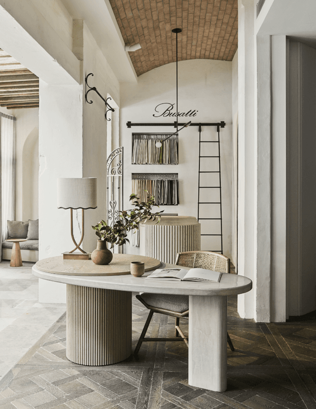 A refined interior vignette featuring a sculptural oval stone or concrete table with cylindrical fluted pedestal bases, a textured table lamp, vase with greenery, and an open book. The space has white plastered walls, arched openings, a curved exposed brick ceiling, dark herringbone flooring, wrought-iron wall hooks, a ladder-style vertical feature, and a ribbed console in the background. The setting reads as a stylish entryway or living area with Mediterranean-inspired architectural detailing and warm neutral finishes.