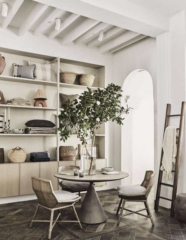 A refined, neutral-toned dining area with a round pedestal table and woven upholstered chairs, set against full-height built-in shelving filled with baskets, cushions, ceramics, and textiles. The room features a white painted exposed-beam ceiling, white walls, a light timber built-in cabinet base, dark patterned timber flooring, an arched wall opening, and a leaning timber ladder with towels. A leafy indoor tree adds an organic element.