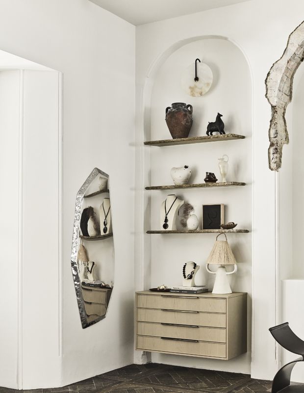 A refined built-in display niche with arched plaster detailing, floating rustic shelves, a light wood drawer cabinet, sculptural decor, jewellery displays, a table lamp, and an irregular antiqued mirror. The space has white walls and dark patterned timber flooring, creating a curated, gallery-like interior.