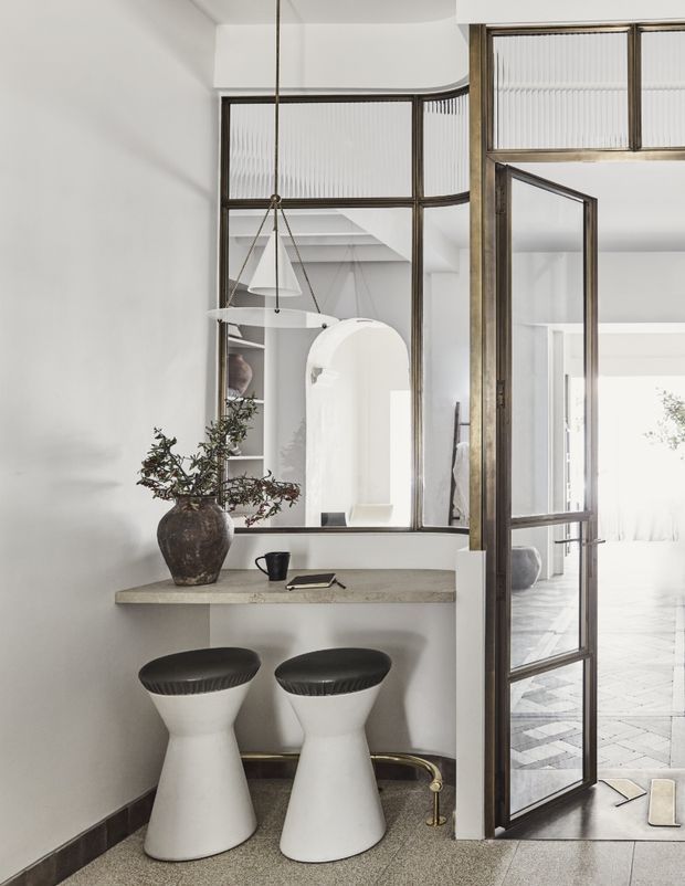 Refined interior vignette showing a compact built-in stone-look console or bar ledge beneath a large internal steel-framed glazed partition. Two sculptural white stools sit below the ledge, with a dark ceramic vase and branch arrangement above. A tall glazed door opens toward a bright adjoining space with patterned flooring. The palette is predominantly white, grey, black, and aged bronze, with strong natural light and elegant industrial detailing.