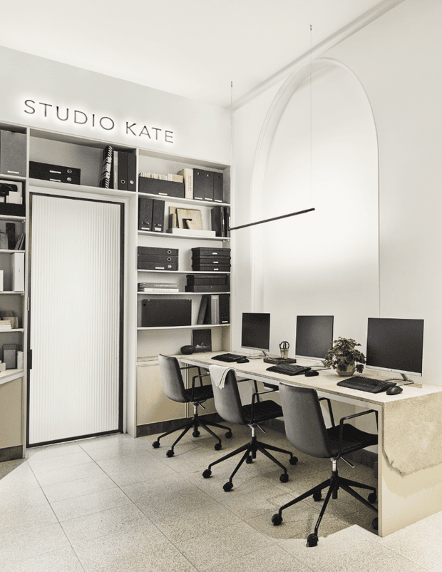 Bright, minimalist studio office with three computer workstations arranged along a long built-in desk. Floor-to-ceiling shelving stores binders and office supplies, while a tall ribbed sliding door and large arched wall detail define the space. The palette is predominantly white, cream, light grey, and black, with tiled flooring and understated contemporary furnishings.