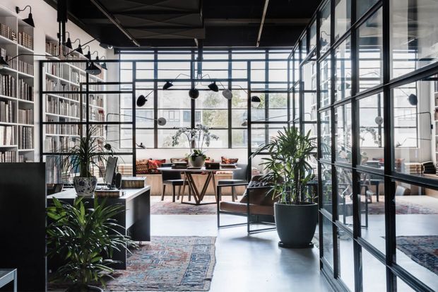 A spacious, light-filled contemporary office or study with floor-to-ceiling black-framed glazed partitions, extensive built-in bookshelves, a desk and work area, lounge seating, indoor plants, patterned rugs, and a striking exposed dark ceiling structure. The open-plan workspace combines industrial metal-and-glass detailing with warm timber furniture and abundant daylight.