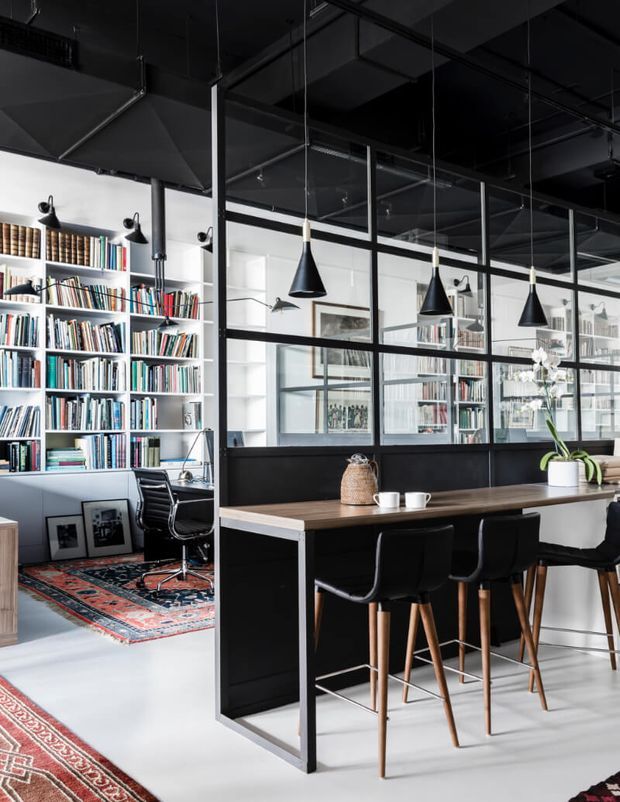 A stylish open-plan interior combining a home office and dining or breakfast bar area. The space features extensive white built-in bookshelves, a black-framed glazed partition, black cabinetry, a long timber-topped counter with bar stools, black pendant lights, exposed dark ceiling services, and patterned rugs. The look is refined and industrial with strong black-and-white contrast and warm timber accents.