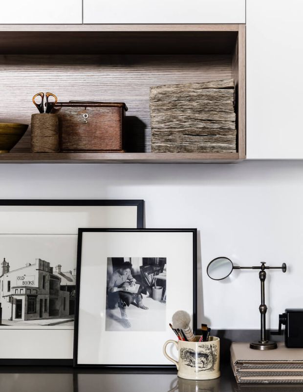 Close-up of a carefully styled home office or study nook featuring open timber shelving with stationery and storage boxes, framed black-and-white artwork leaning against a white wall, a desk lamp, and a ceramic cup holding brushes or pens. The composition has a refined, monochromatic palette with warm wood accents and layered decorative objects.