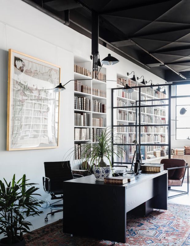A refined home office or study with a black desk, office chair, extensive built-in bookshelves, black-framed glazed partitions, pendant and wall lighting, indoor plants, and a patterned area rug. The room combines crisp white walls with dark industrial ceiling elements and abundant natural light.