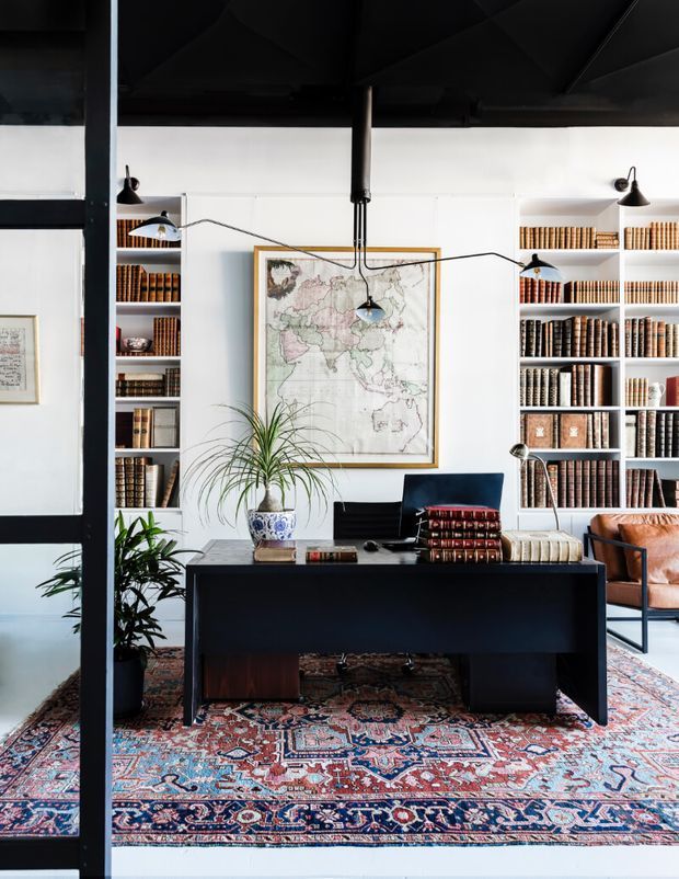 A refined home office or study with a large black desk centered before built-in white bookshelves filled with leather-bound books. A framed antique-style world map hangs behind the desk, complemented by articulated black task lighting, potted plants, a laptop, and layered Persian-style rugs. A tan leather armchair is visible to the right. The room combines a dark ceiling and structural framing with bright white walls and classic scholarly furnishings.