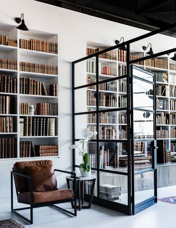 A refined library or home office with floor-to-ceiling white built-in bookshelves densely filled with leather-bound books. Black-framed glass partition doors divide the space, while a brown leather lounge chair, small side table, black wall sconces, and a potted orchid create a sophisticated reading area. The room has a bright, high-contrast palette with industrial-style metal detailing and polished light flooring.
