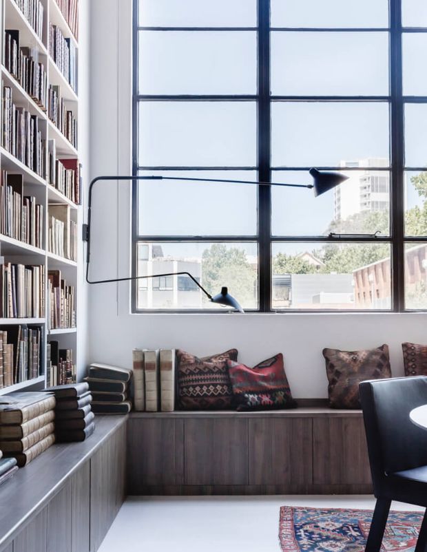 A bright, tailored reading nook or library corner with floor-to-ceiling built-in bookshelves, a dark timber built-in window seat and cabinetry, patterned cushions, a large black-framed industrial-style window, and a slim black articulated wall lamp. A partial dark armchair and patterned rug are visible in the foreground. The space has a refined contemporary character with strong built-in joinery and abundant daylight.