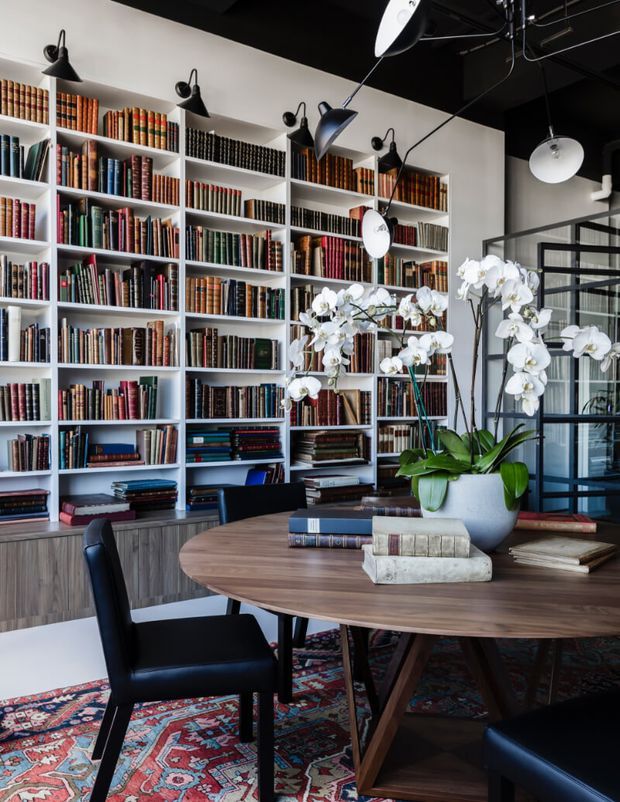A sophisticated library and dining workspace with full-height white built-in bookshelves filled with books, a large round timber dining table, black leather chairs, and a vivid patterned rug. Black-painted ceiling services and sculptural pendant lighting create a dramatic contrast, while orchids and glazed steel-framed partitions add refinement.