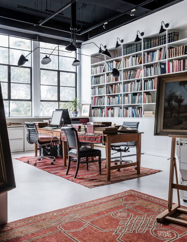 A spacious, eclectic office or library with full-height white bookshelves filled with books, large black-framed industrial-style windows, exposed black ceiling services, and multiple black articulated pendant and wall lights. A timber worktable with office chairs occupies the centre, layered red patterned rugs add warmth, and framed artwork is displayed on an easel and wall.