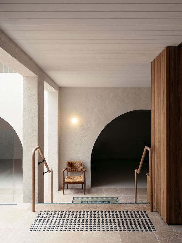 Interior architectural view of a refined transitional space with a stair or level change in the foreground. The room features pale textured plaster walls, a white horizontal-panel ceiling, pale stone or porcelain flooring, timber handrails, a dark timber slatted screen or joinery wall, and a large rounded arch opening leading into a dark adjoining area. A woven wooden chair and minimal wall light add warmth. The composition emphasizes tactile materials, strong geometric forms, and high natural light from the left.