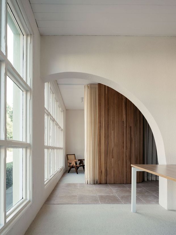 A bright, narrow interior passage or living area with full-height multi-pane windows along one side, pale carpet in the foreground, large beige stone-look tiled flooring beyond, and a broad white curved arch opening. The arch frames a timber-lined feature wall and concealed doorway, with sheer curtains and two wooden lounge chairs at the far end. A simple light timber-topped table or desk occupies the right foreground. The space has clean, restrained detailing and strong natural light.