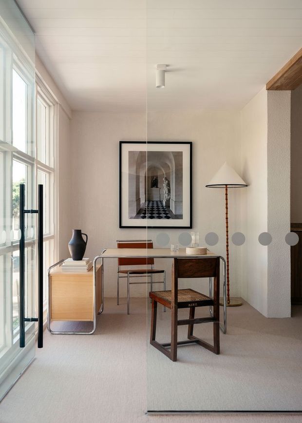 Bright, refined home office or study with a minimalist desk positioned beside tall windows and glazing. The room features pale walls and carpet, a warm timber sideboard, dark timber chair, framed architectural artwork, a slender floor lamp, and a partial timber ceiling beam or trim at the right. The space has a calm, contemporary character with subtle mid-century-inspired furniture and strong natural light.
