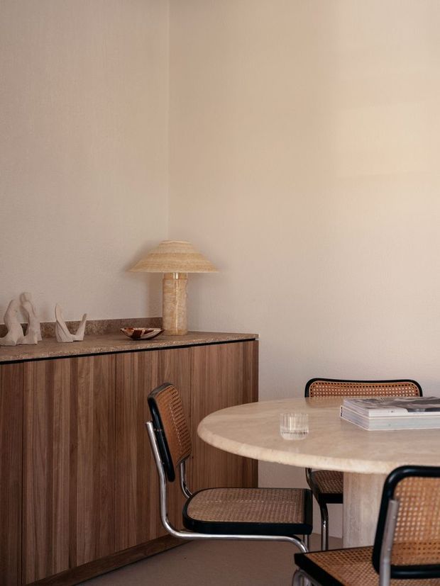 Warm, softly lit dining area with a round stone-topped table and woven cane dining chairs. A built-in dark timber sideboard with a dark stone benchtop lines the wall, styled with a table lamp and small sculptural objects. The space has a restrained, refined contemporary feel with neutral plaster walls and rich wood tones.