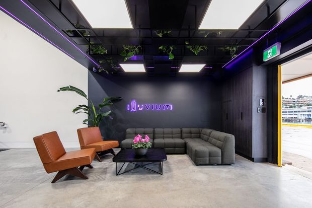 A contemporary commercial lounge or reception area with a large grey sectional sofa, two rust-coloured leather lounge chairs, and a black geometric coffee table. The space features a dark feature wall with illuminated purple signage, black ceiling panels with integrated linear and square lighting, hanging plants, polished concrete flooring, and an open glazed doorway to the exterior.
