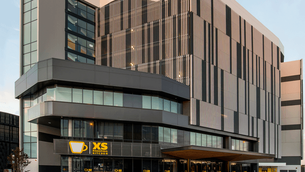 Exterior view of a contemporary multi-storey commercial building with a dark grey and light facade. The main elevation features extensive glazing, projecting horizontal volumes, and tall vertical timber-toned fins forming a screened facade. Ground-level retail or hospitality frontage includes prominent signage and a broad timber-toned canopy.