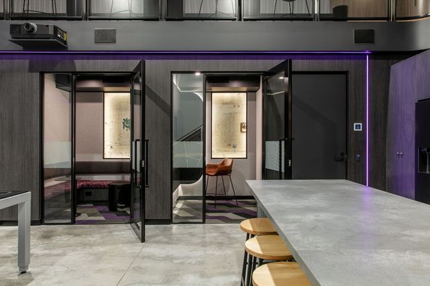 Contemporary commercial interior with a large concrete-look island or counter in the foreground, timber stools, dark charcoal wall finishes, black-framed glazed rooms, and purple linear accent lighting. The glass-enclosed spaces contain lounge and work seating, with exposed services and an industrial-style ceiling visible above.