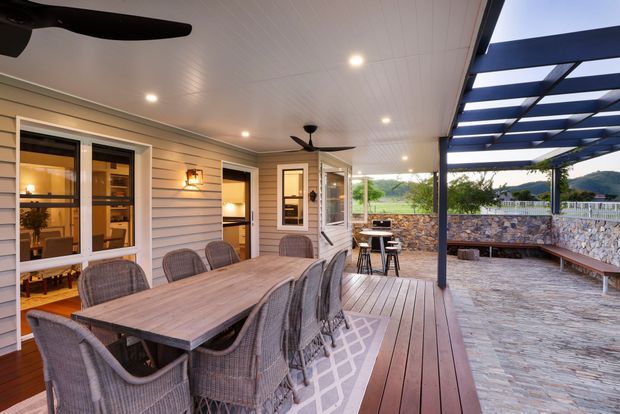 Covered outdoor entertaining area with a large timber dining table and woven chairs on a timber deck. The space features a slatted pergola, ceiling fans, recessed lighting, light horizontal weatherboard walls, multiple glazed doors and windows, an outdoor cooking or bar area, and a stone-paved courtyard with a stone retaining wall beyond.